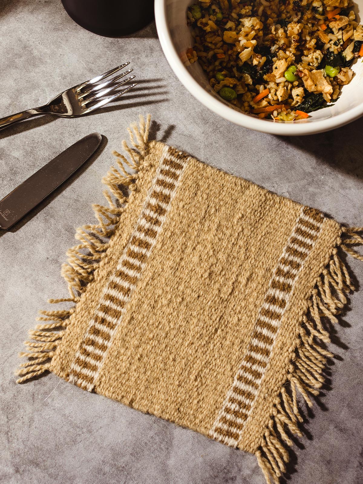 Handwoven beige trivet with fringe edges on a grey stone table, paired with a knife and fork, next to a bowl of mixed vegetables and rice.