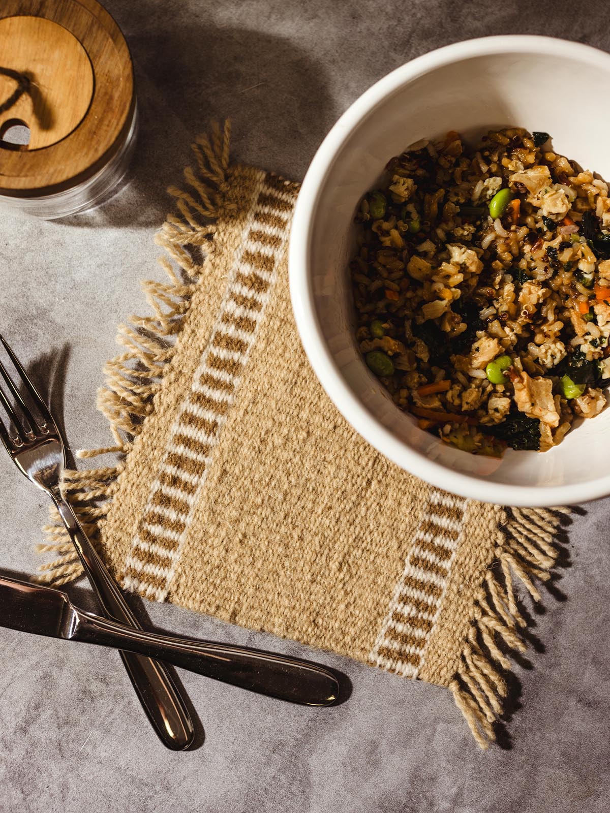 Top view of a handwoven beige trivet with fringe edges placed on a grey stone table, featuring a white bowl of mixed vegetables and rice, with a knife and fork set on the side.