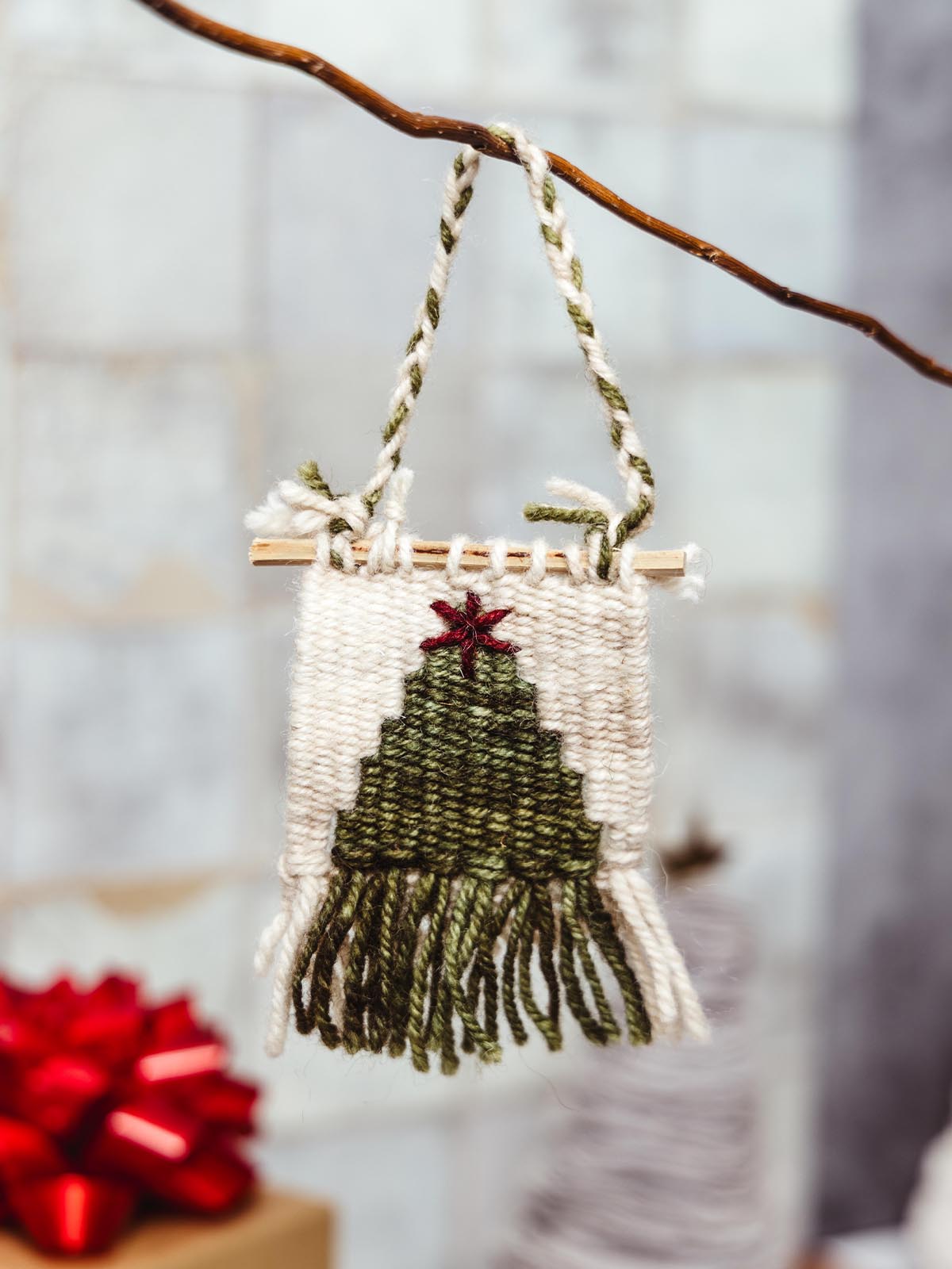 Handwoven holiday ornament featuring a green tree design with fringe, hanging on a branch. Artisan-crafted wall hanging for festive home decor.