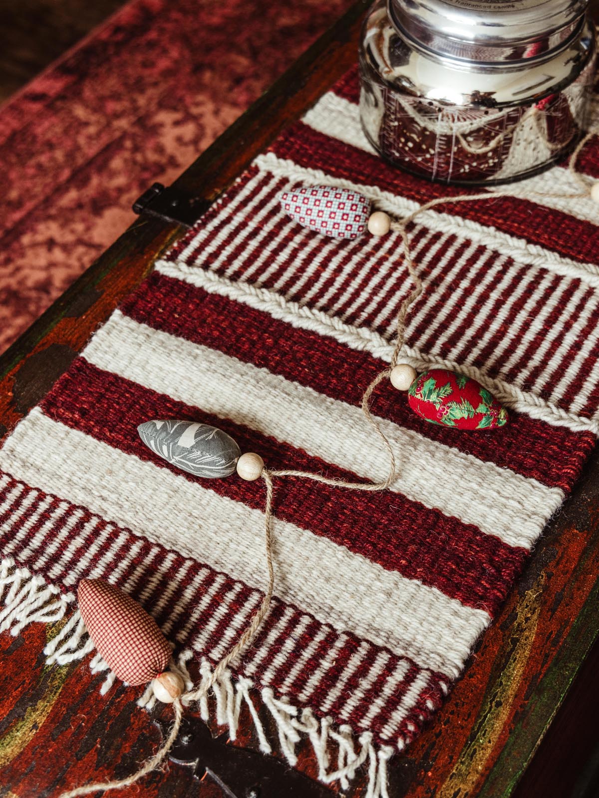 Decorative fabric garland with wooden beads placed on a red and white striped woven table runner. Handcrafted holiday decor on a rustic wooden chest, adding festive charm to home interiors.