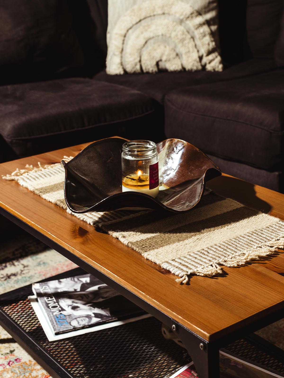 A black ceramic bowl with a candle placed inside, sitting on a beige and brown striped woven table runner, displayed on a wooden coffee table with a dark sofa in the background.