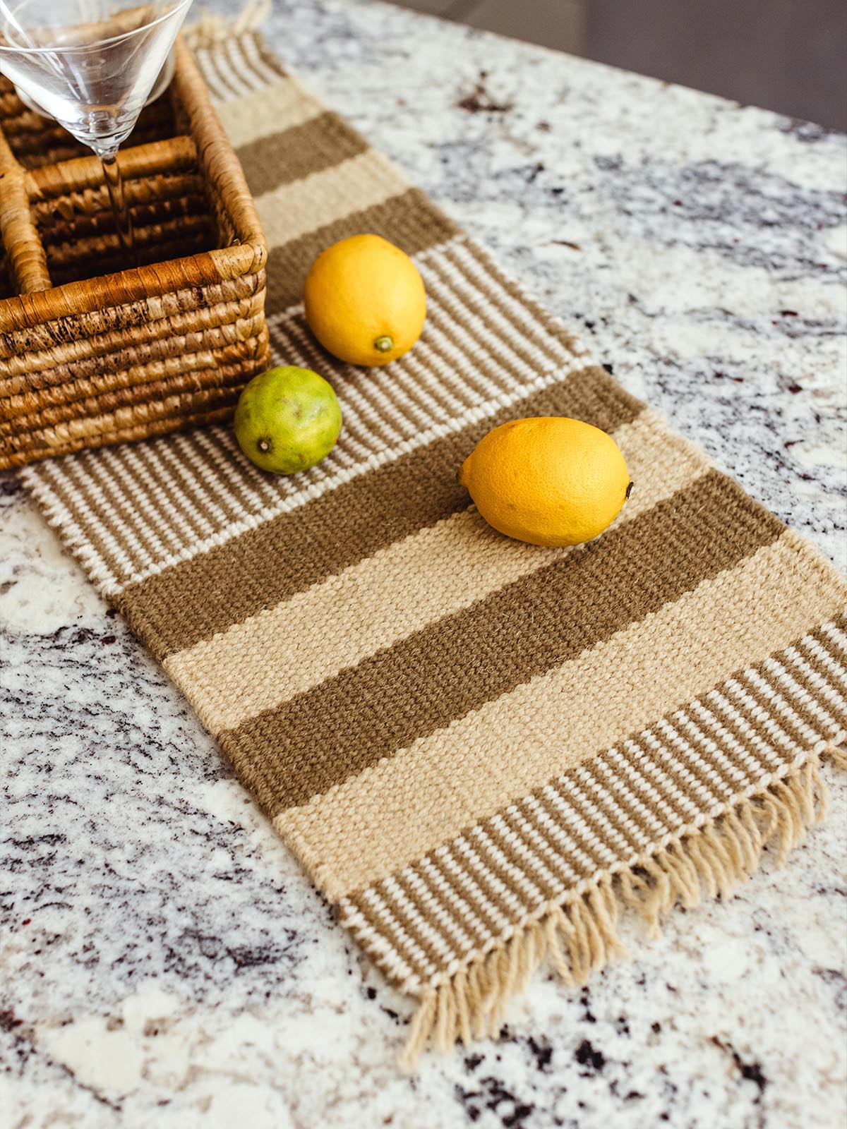 Close-up of a beige and brown striped woven table runner on a granite countertop, adorned with lemons and limes, next to a woven basket holding glassware.