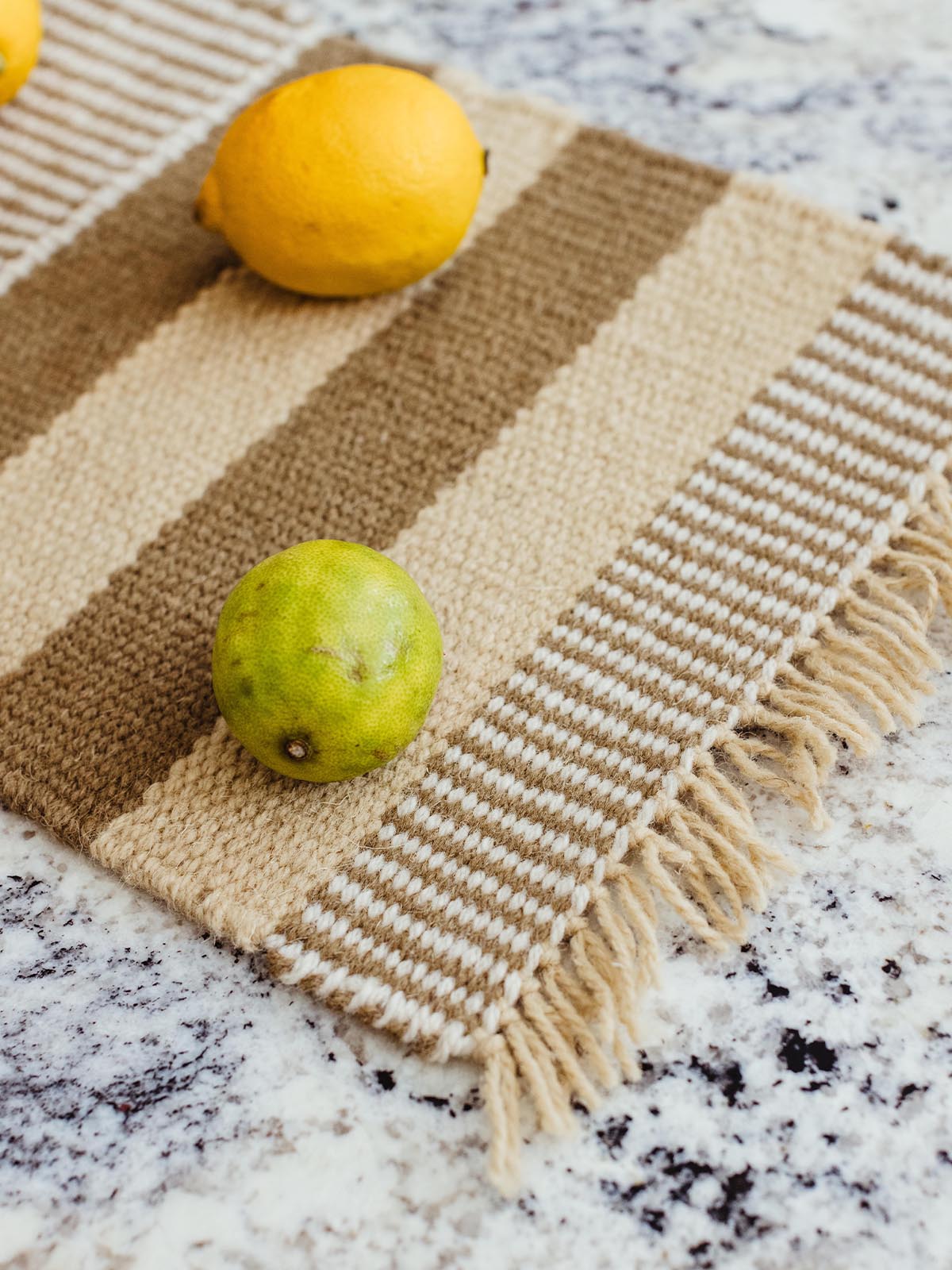 Beige and brown striped woven table runner with lemons and limes scattered on it, placed on a speckled granite countertop.