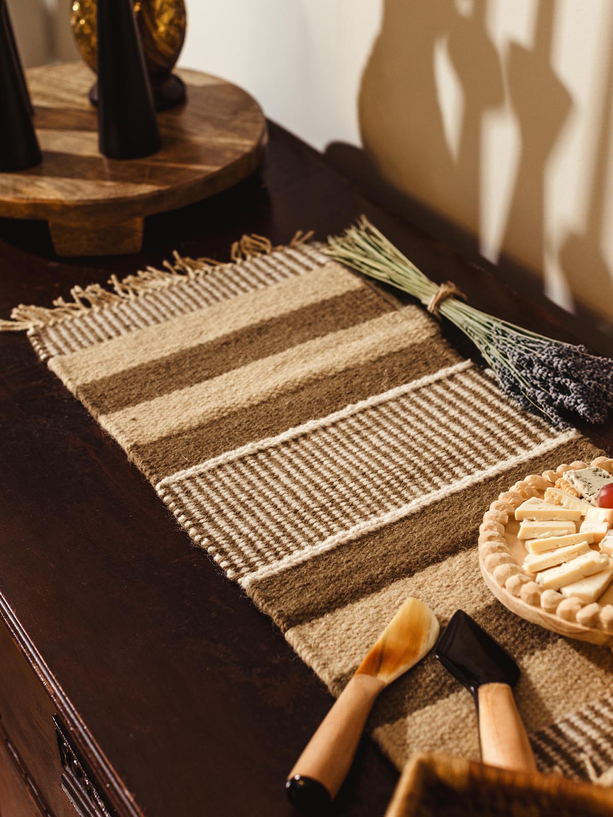 Beige and brown striped woven table runner displayed on a dark wooden sideboard, decorated with lavender, a cheese platter, and wooden spreaders.