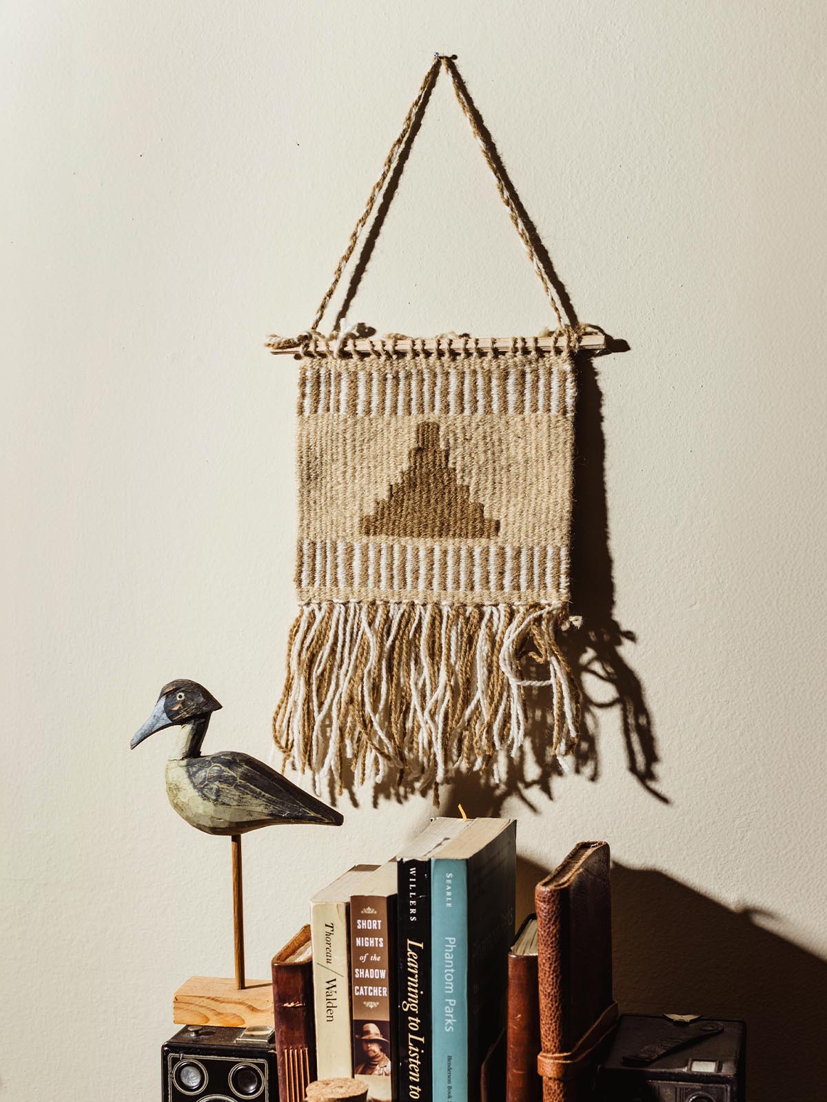 Small woven wall hanging with a geometric pattern in beige and brown, displayed on a cream-colored wall above a shelf holding a wooden bird figurine and a stack of vintage books.