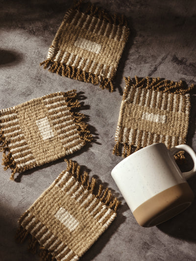 Set of four handwoven coasters with earthy tones, arranged on a rustic surface alongside a ceramic mug.